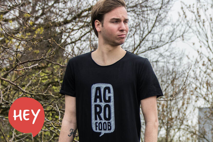 Man met een zwart shirt met de tekst 'Acrofoob' erop staat in een tuinrijke omgeving