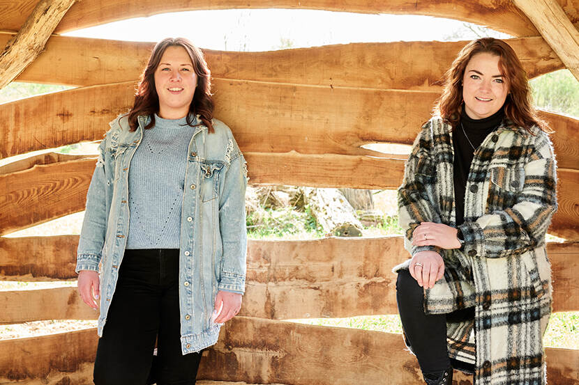 Nicky en Lieke, twee jonge vrouwen, staan voor een houten achtergrond en kijken naar de camera